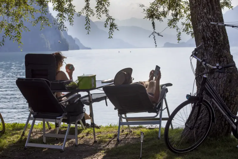 Two guests in camping chairs under a tree by the lake at Camping Aaregg